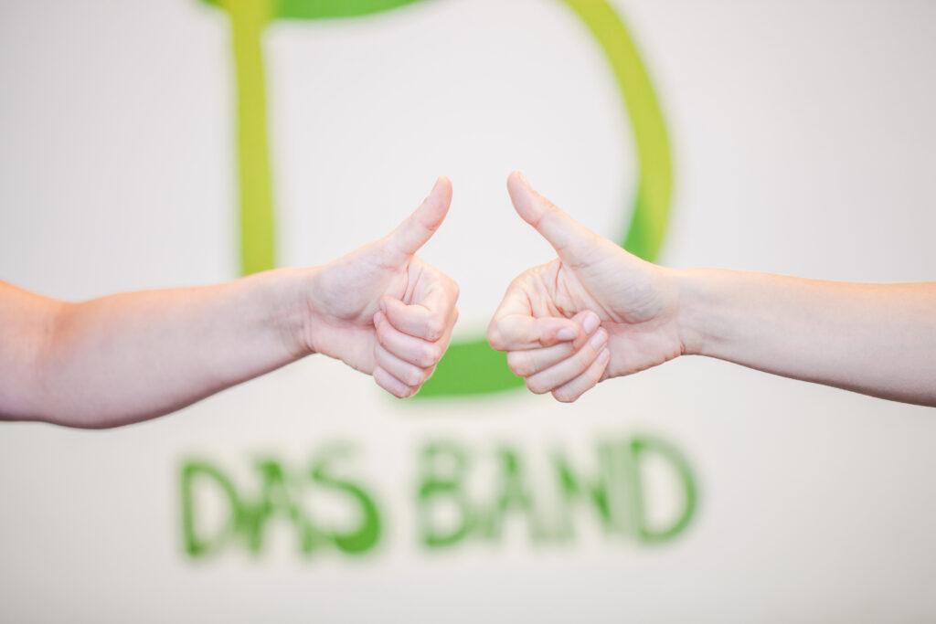 Hände mit Daumen hoch vor einer Wand mit Schriftzug DAS BAND